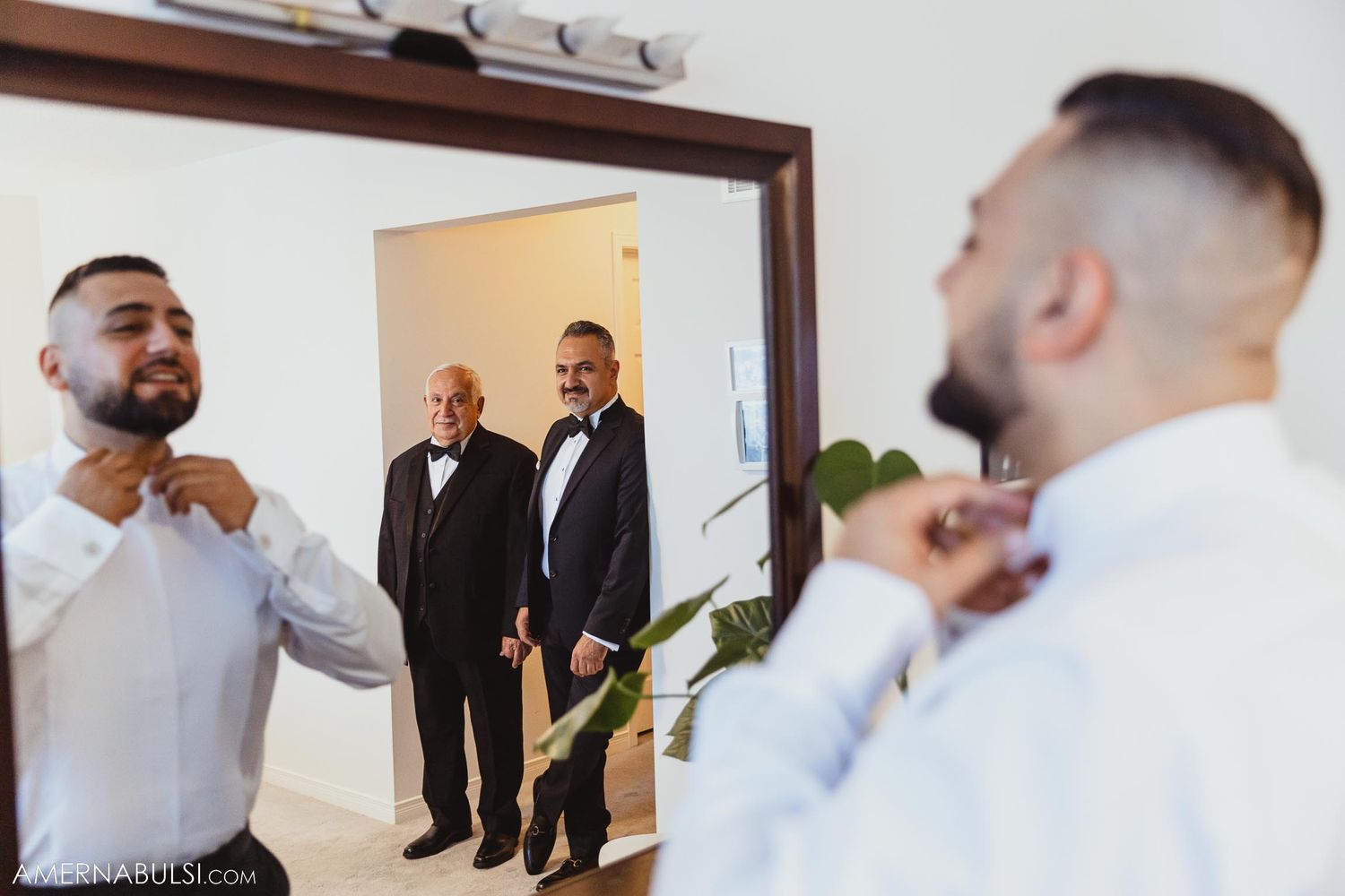 A person adjusts their tie in a mirror while others look on during wedding preparations.