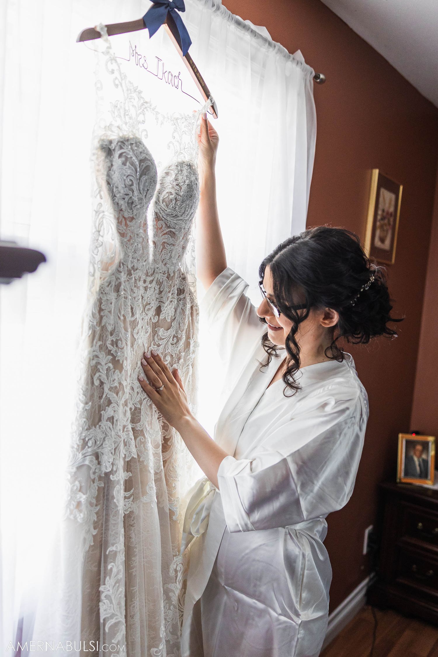 An intricate white lace wedding gown displayed on a hanger against sheer curtains.
