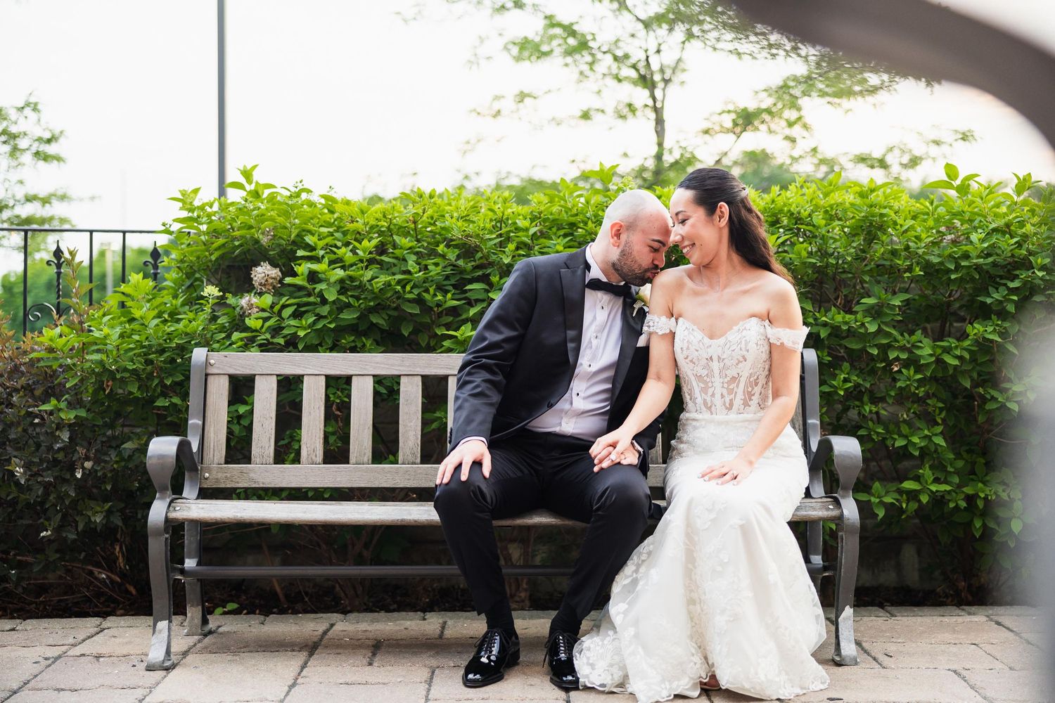 A couple shares a tender moment while sitting together on a garden bench surrounded by greenery.