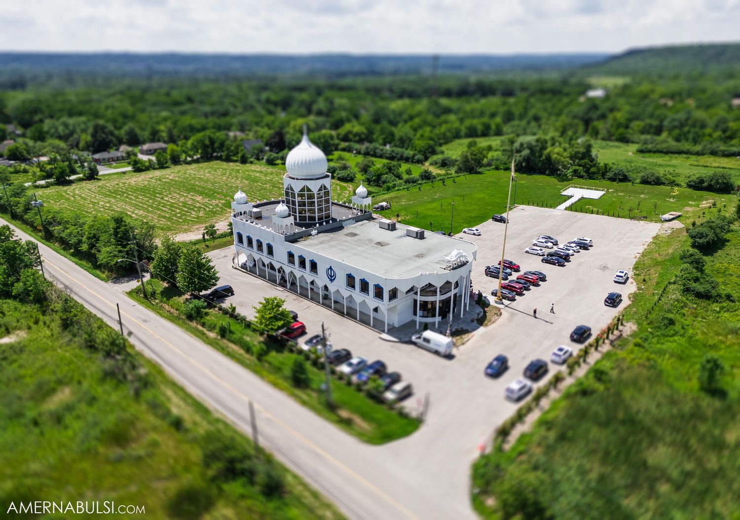 Aerial view of a white Sikh Gurdwara temple with dome and parking lot surrounded by green landscape.