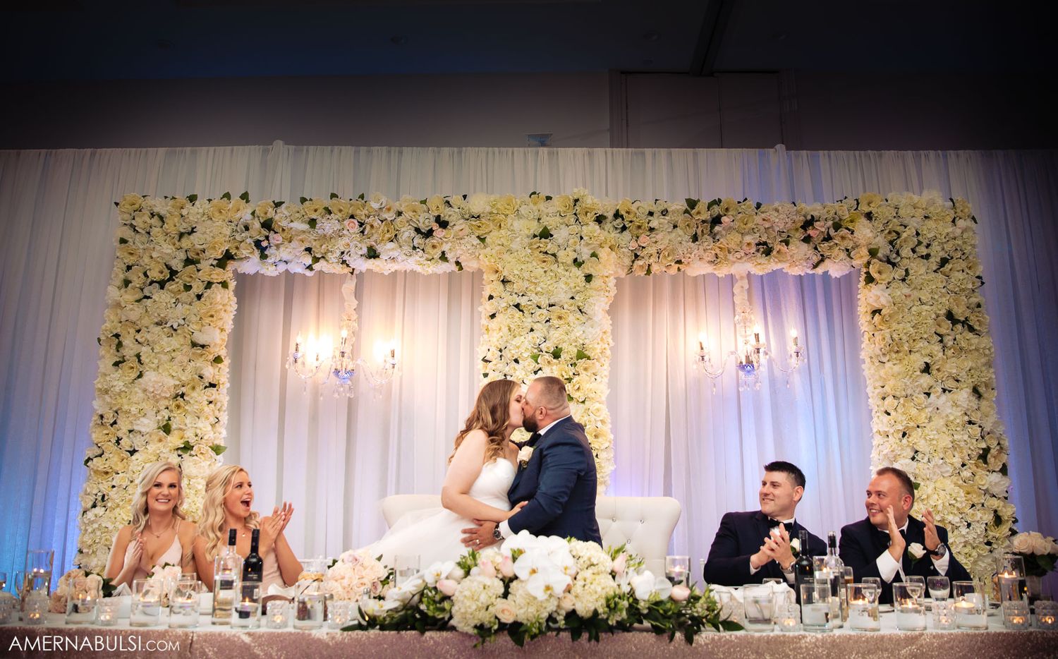 Wedding reception venue with white floral decor and elegant draping along a head table.