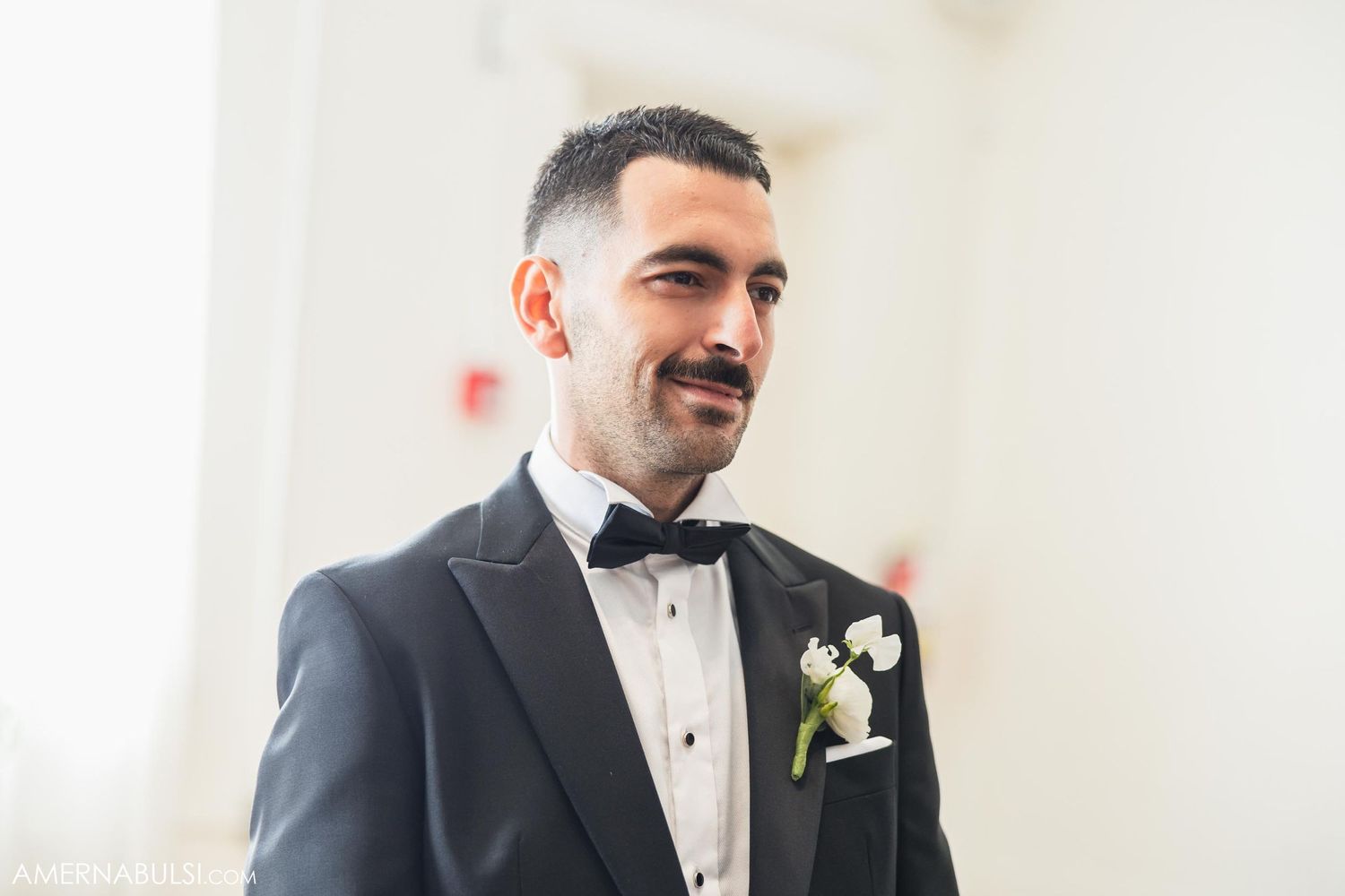 Groom in classic black tuxedo with boutonniere smiles during wedding ceremony.