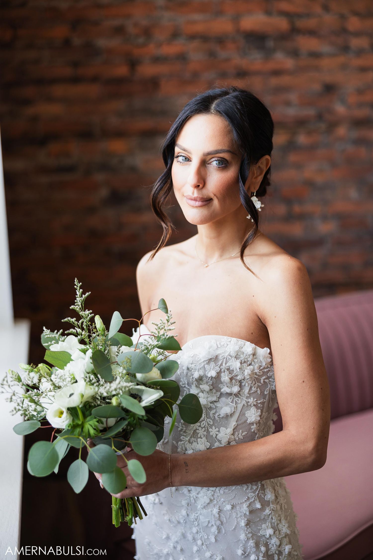 Bride in strapless lace gown holds white and green bouquet against brick wall.