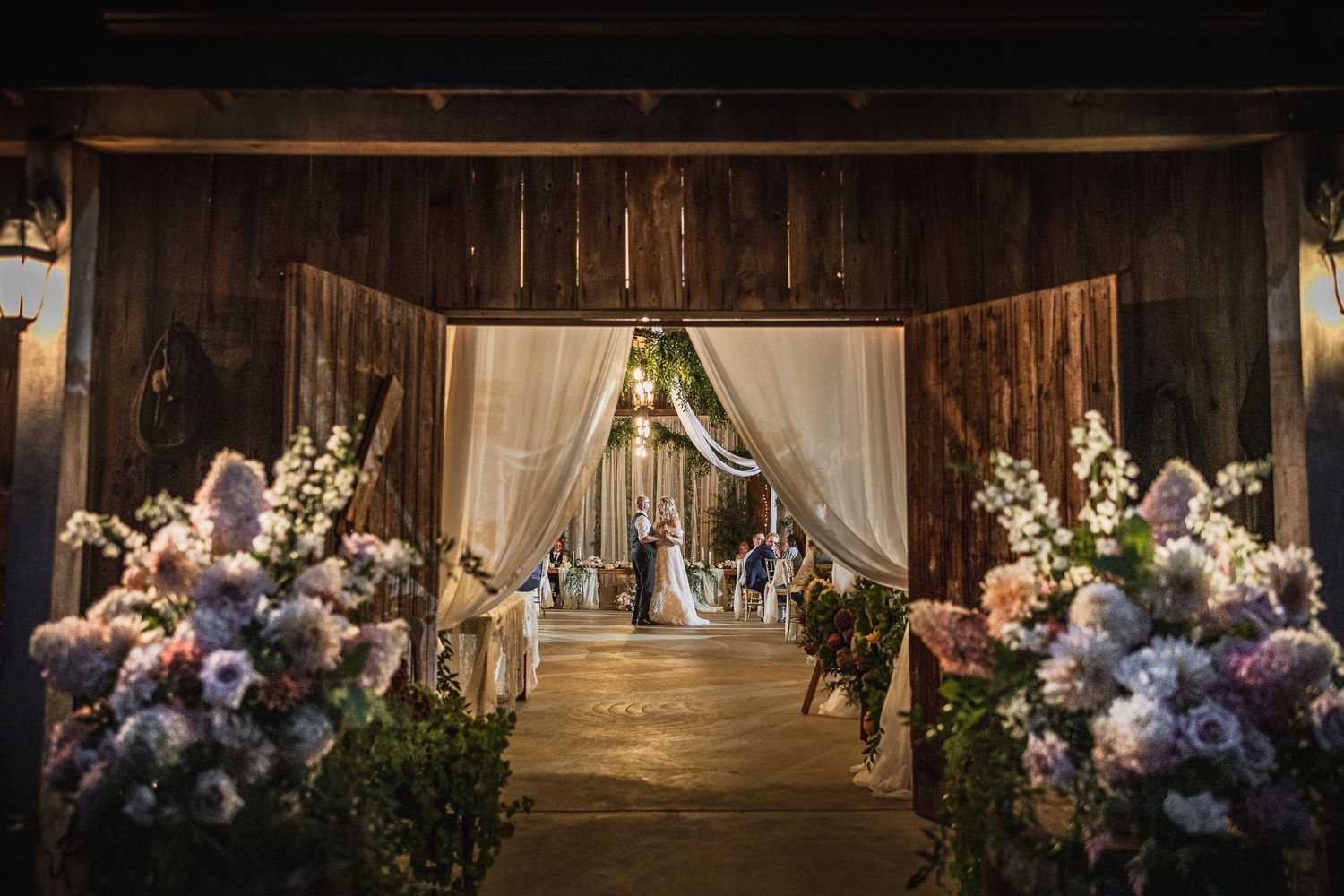 Rustic barn wedding ceremony setup with elegant floral arrangements and draped fabric creating a romantic aisle.