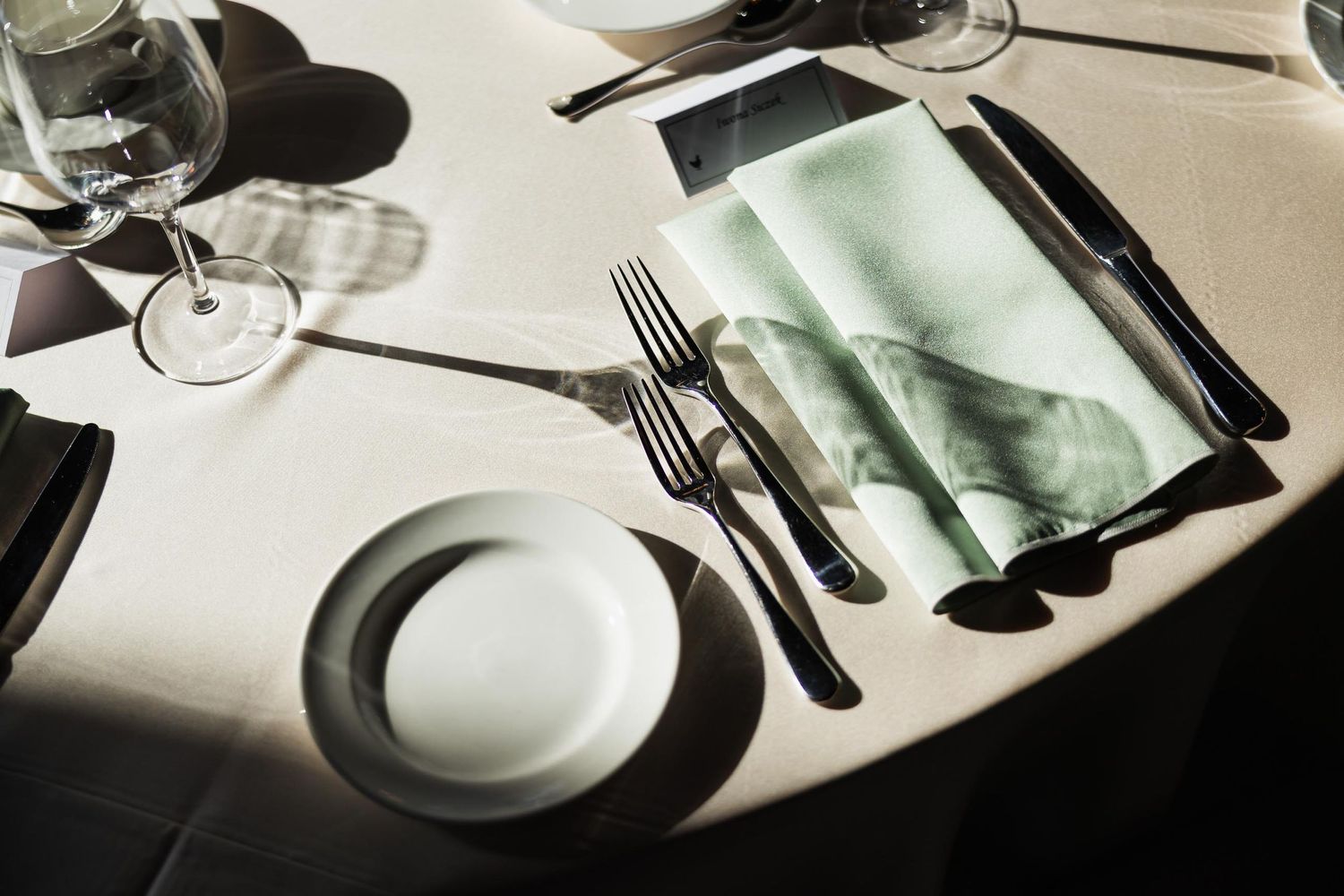 Elegant place setting with silverware, white plate and mint green napkin on a white tablecloth in natural light.
