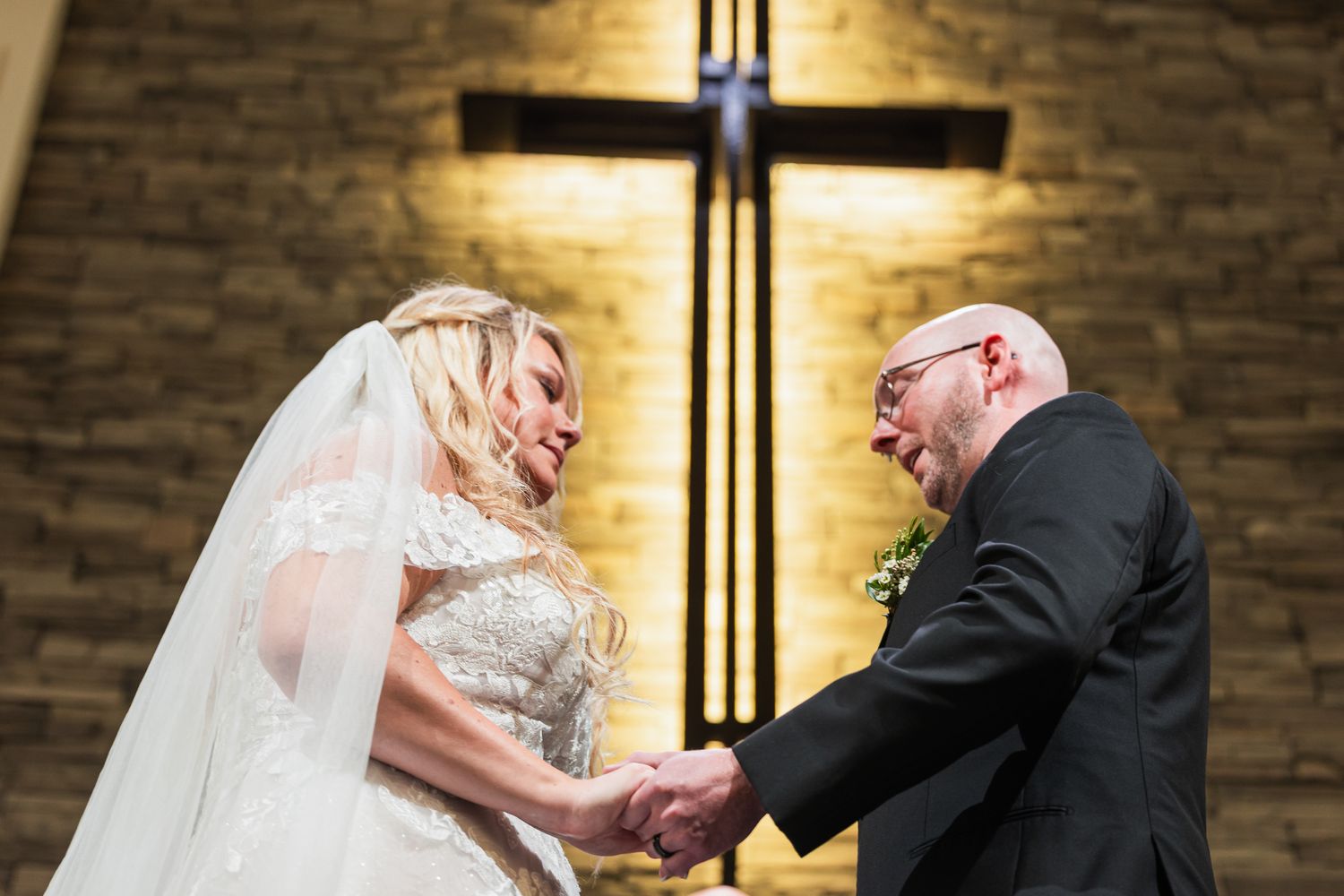 A couple exchanges rings during a church wedding ceremony under warm lighting and stone wall backdrop.