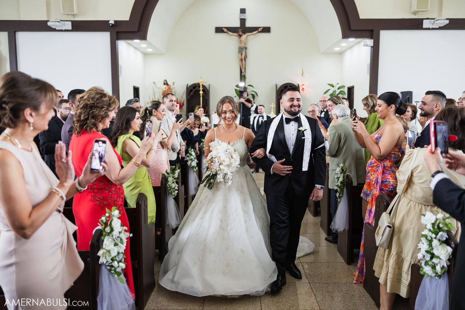 Newlyweds walk down church aisle as wedding guests applaud and take photos.