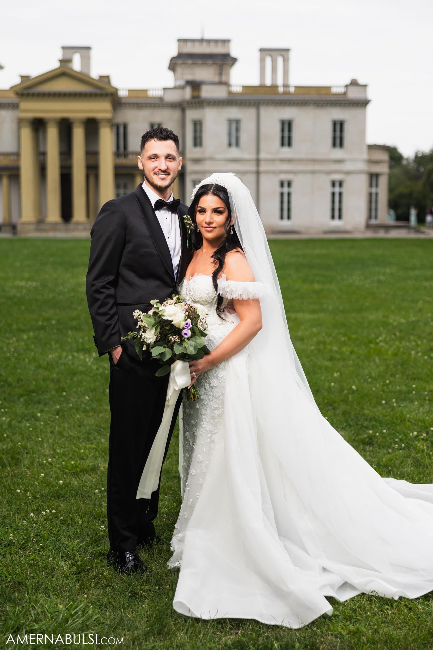 Wedding couple poses in formal attire in front of a grand historic mansion with classic columns.