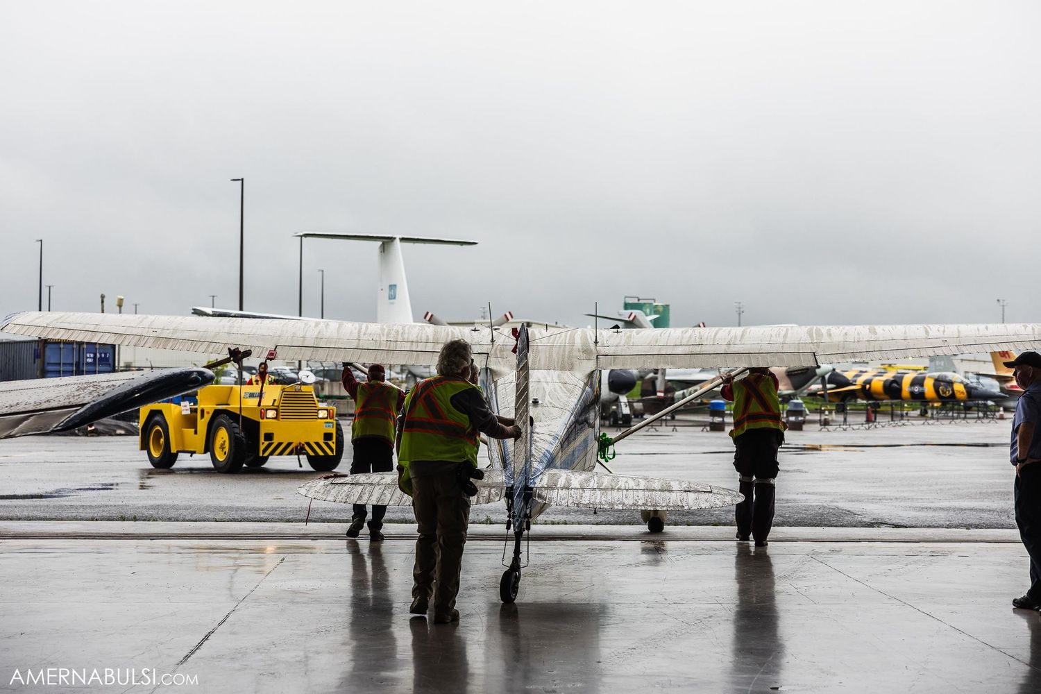 Aircraft maintenance crew inspects a private jet on a rainy tarmac with yellow service vehicle nearby.