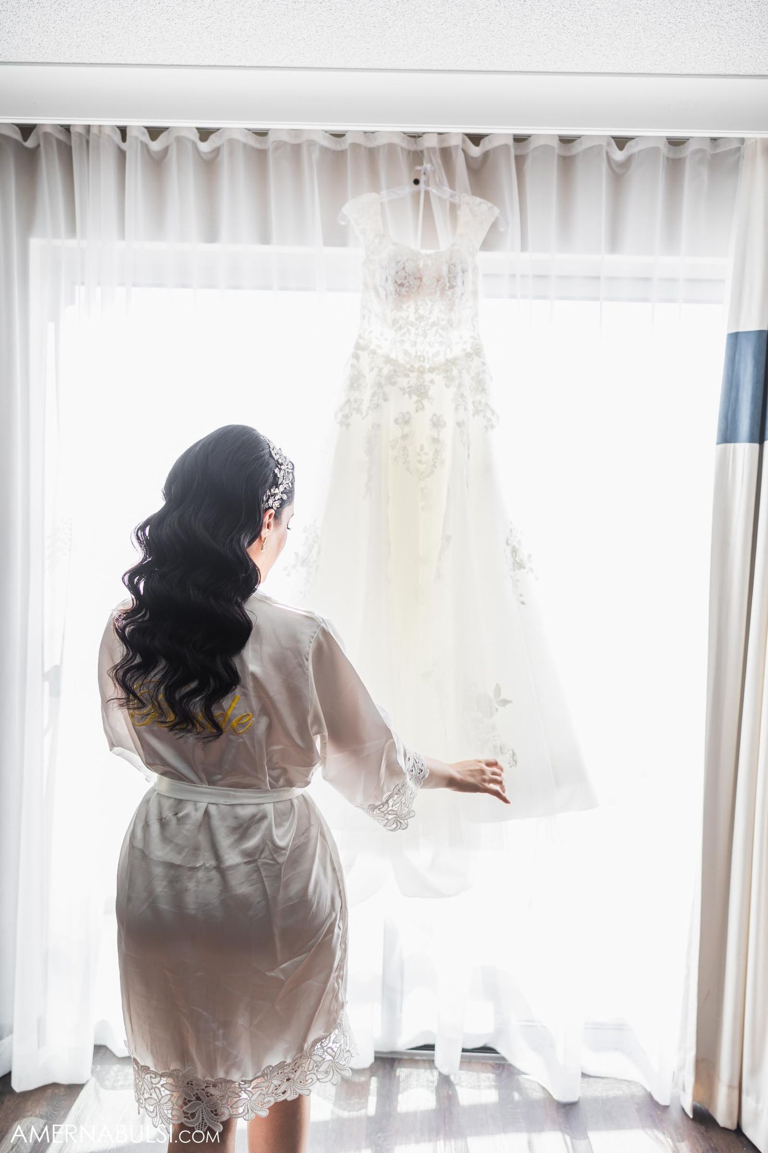 Person in silk robe admiring white wedding dress hanging by sunlit window.