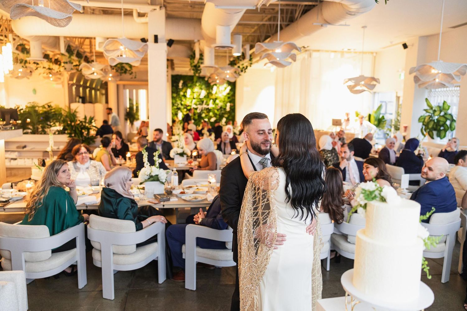 A wedding reception venue decorated with greenery and white chairs as guests dine at tables.
