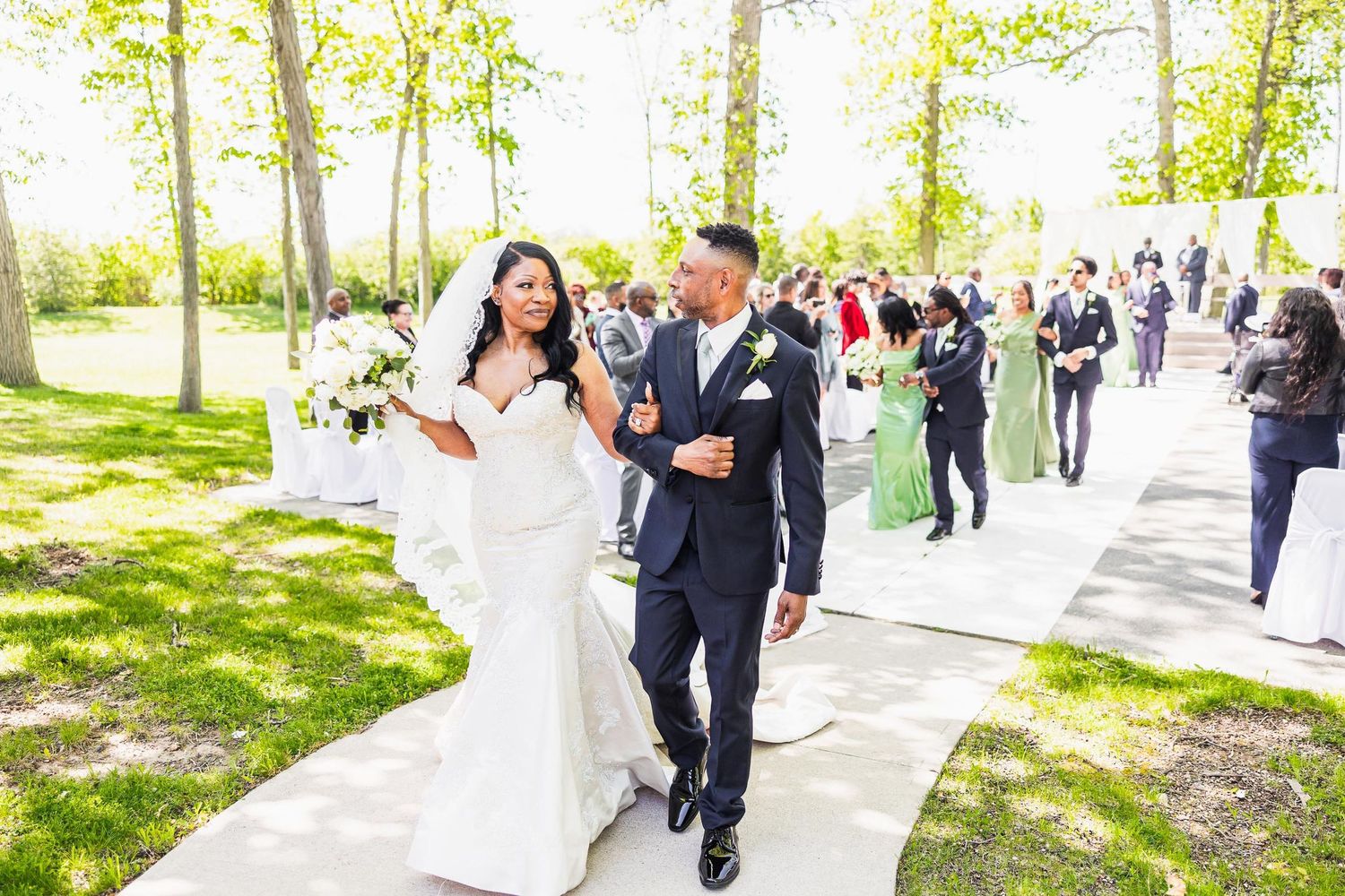 Two people in wedding attire walking together on a tree-lined path surrounded by wedding guests.