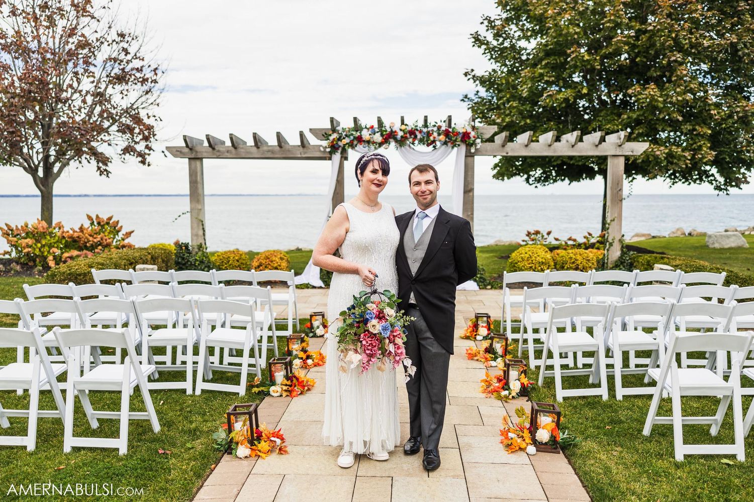 Outdoor wedding ceremony setup with white chairs and fall-colored floral aisle decorations.
