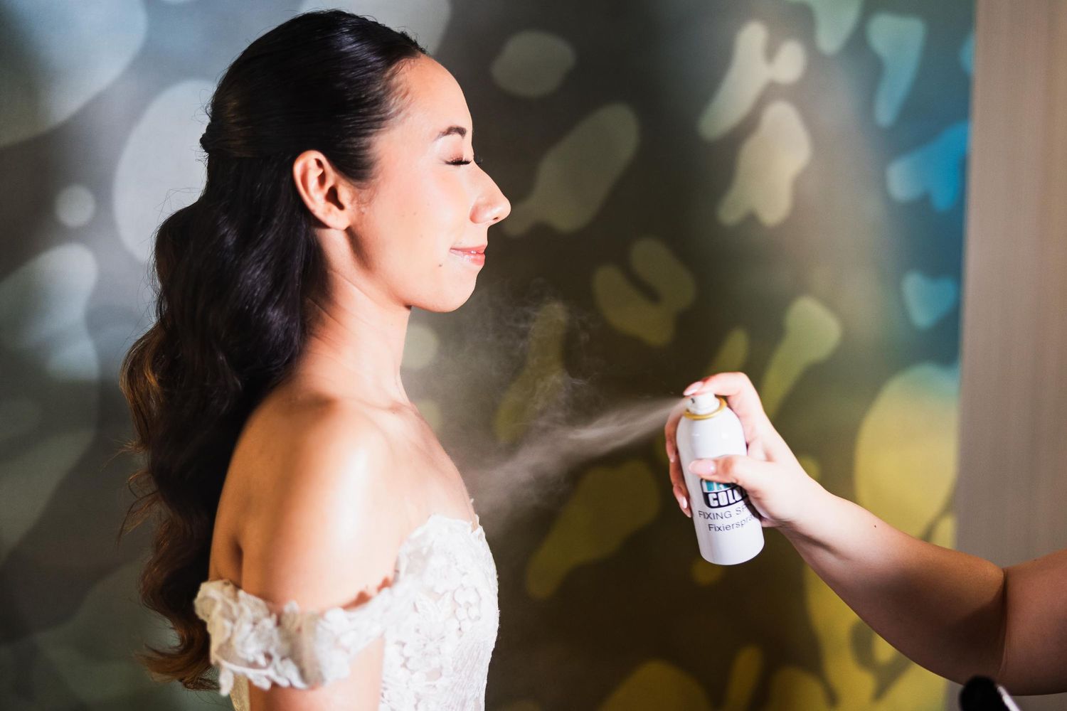 Someone spraying hairspray on a bride wearing an off-shoulder wedding dress against a bokeh background.