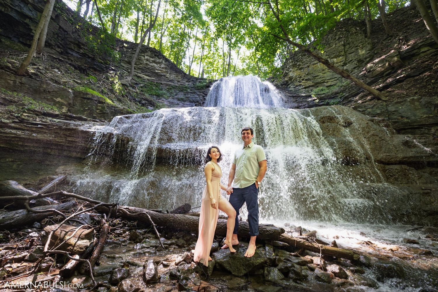 A couple poses together on rocks with a majestic multi-tiered waterfall cascading behind them.