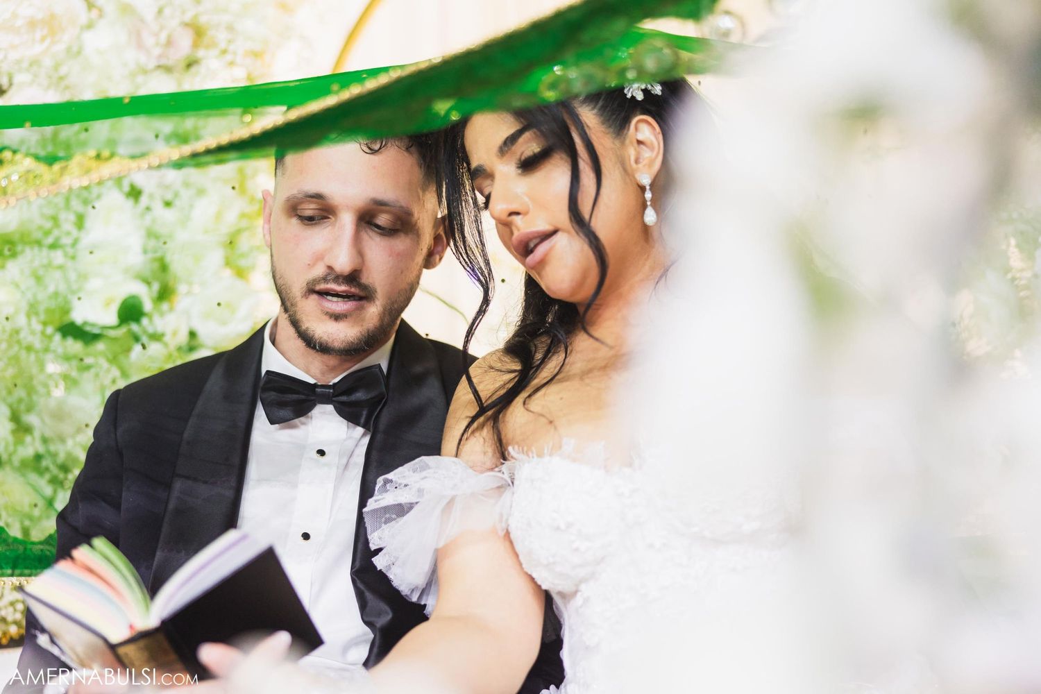 A couple share an intimate outdoor moment while reading together under green foliage.