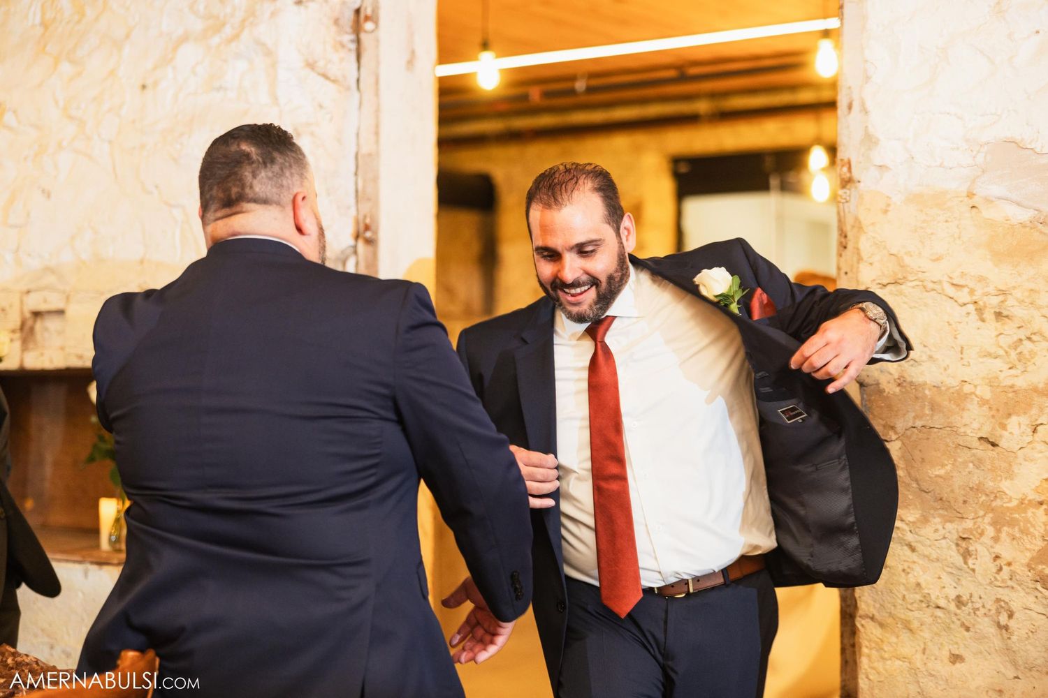 Wedding guests greeting and embracing in a rustic stone-walled hallway.