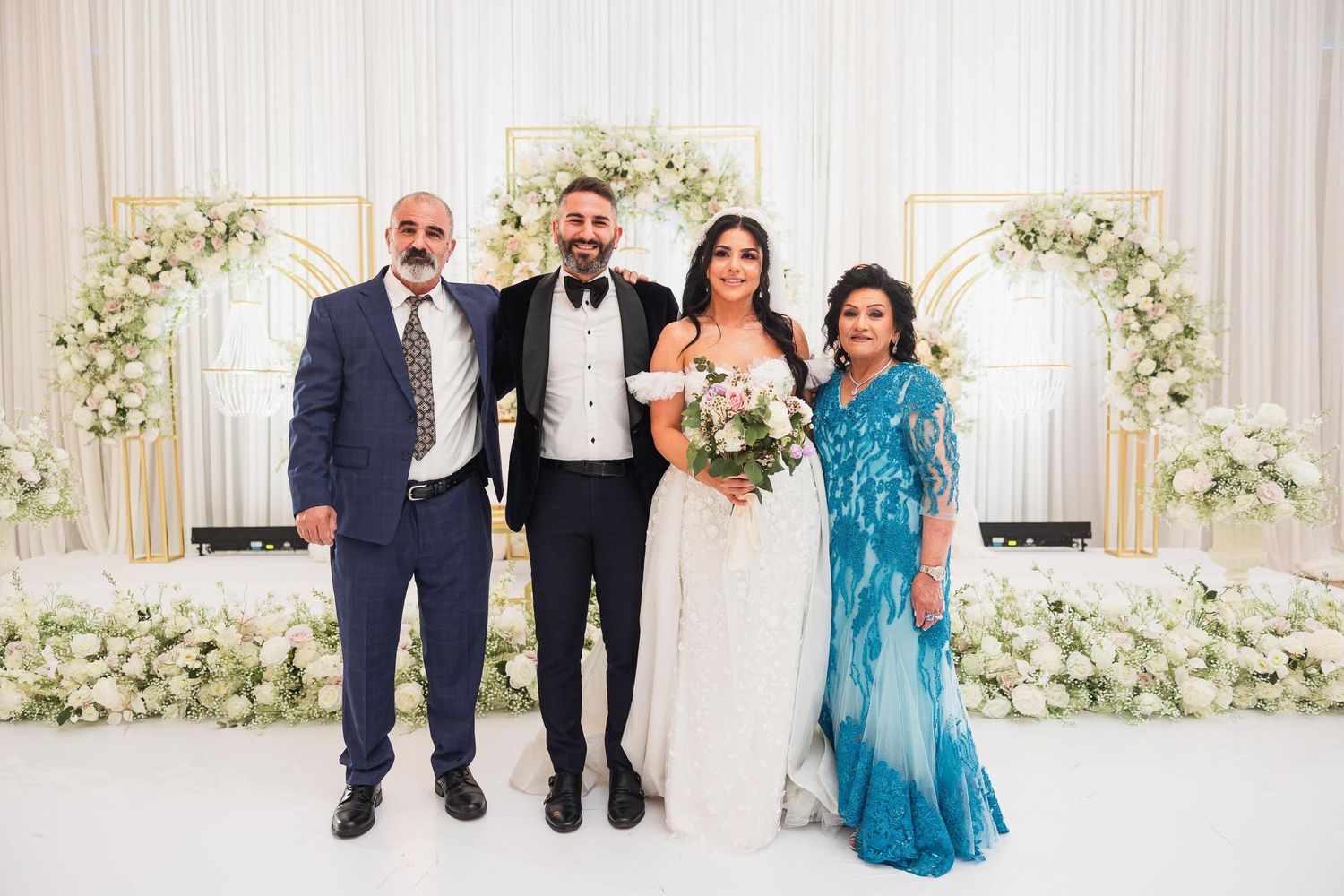 Wedding party poses under elegant white floral arch with draped fabric backdrop.