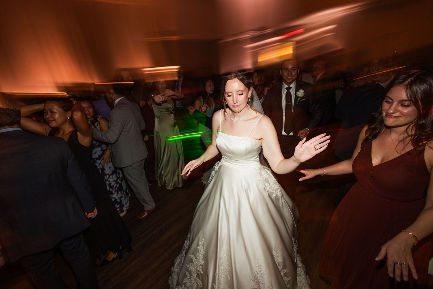 Bride in white ball gown dancing joyfully with guests at a lively wedding reception with colorful lighting.