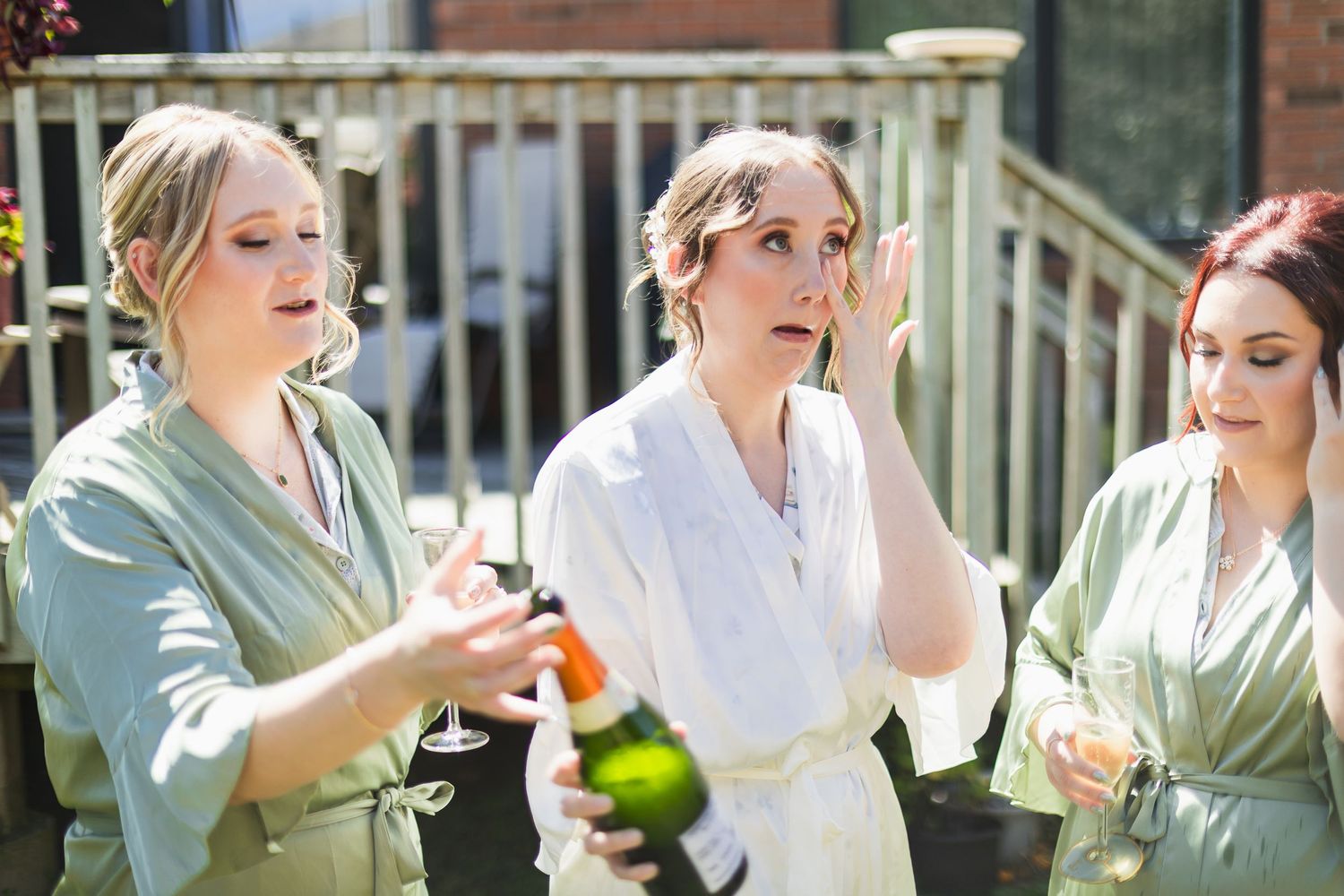 Bride in white robe reacts to champagne bottle with bridesmaids on sunny patio.