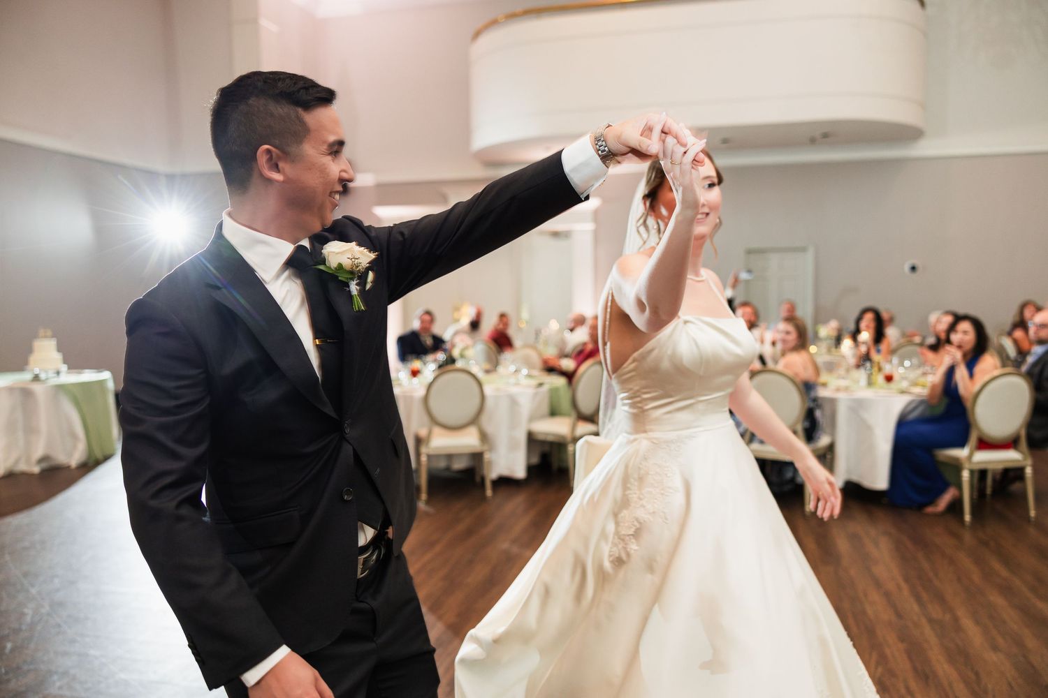 Groom in black tuxedo twirling bride in white gown during elegant wedding reception first dance.