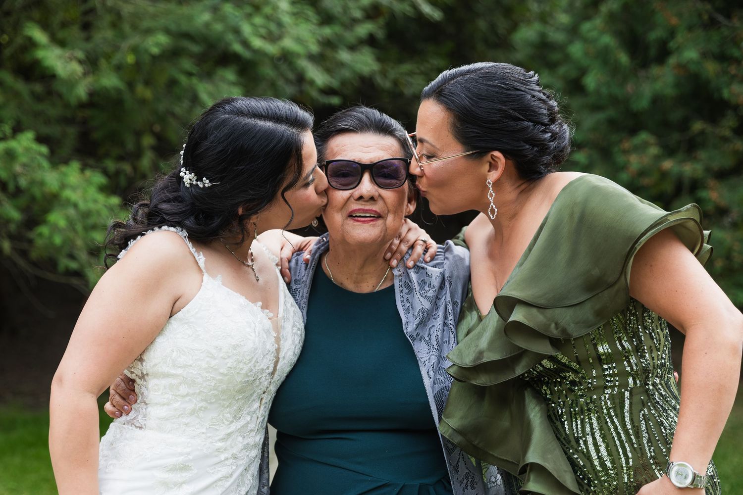 Bride and bridesmaid in green dress kiss the cheeks of a smiling older woman outdoors.