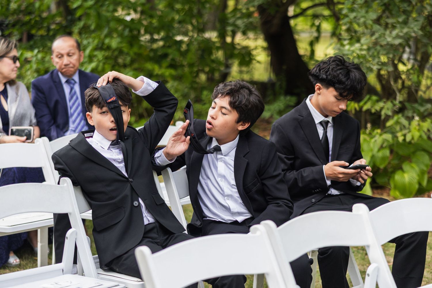 Three young boys in black suits sit in white chairs, distracted on phones during a wedding ceremony.