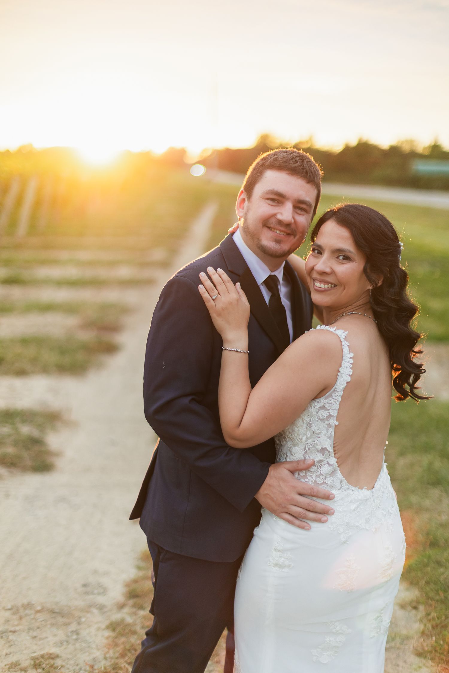 Bride and groom embracing at golden sunset on a rustic path, bride wearing backless lace wedding gown.