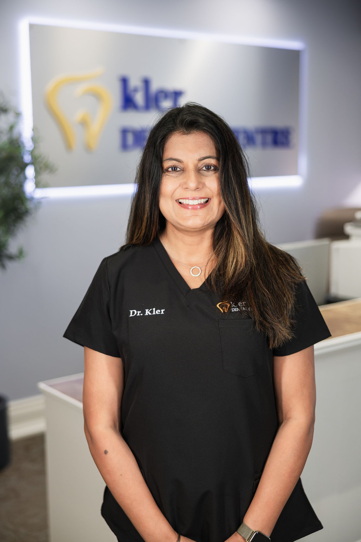 Dr. Kler smiles in black scrubs at Kler Dental Centre reception, showcasing a welcoming dental professional.