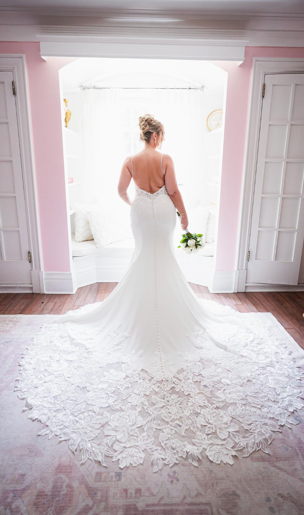 Elegant bridal gown with intricate lace train photographed in a pink-walled room.