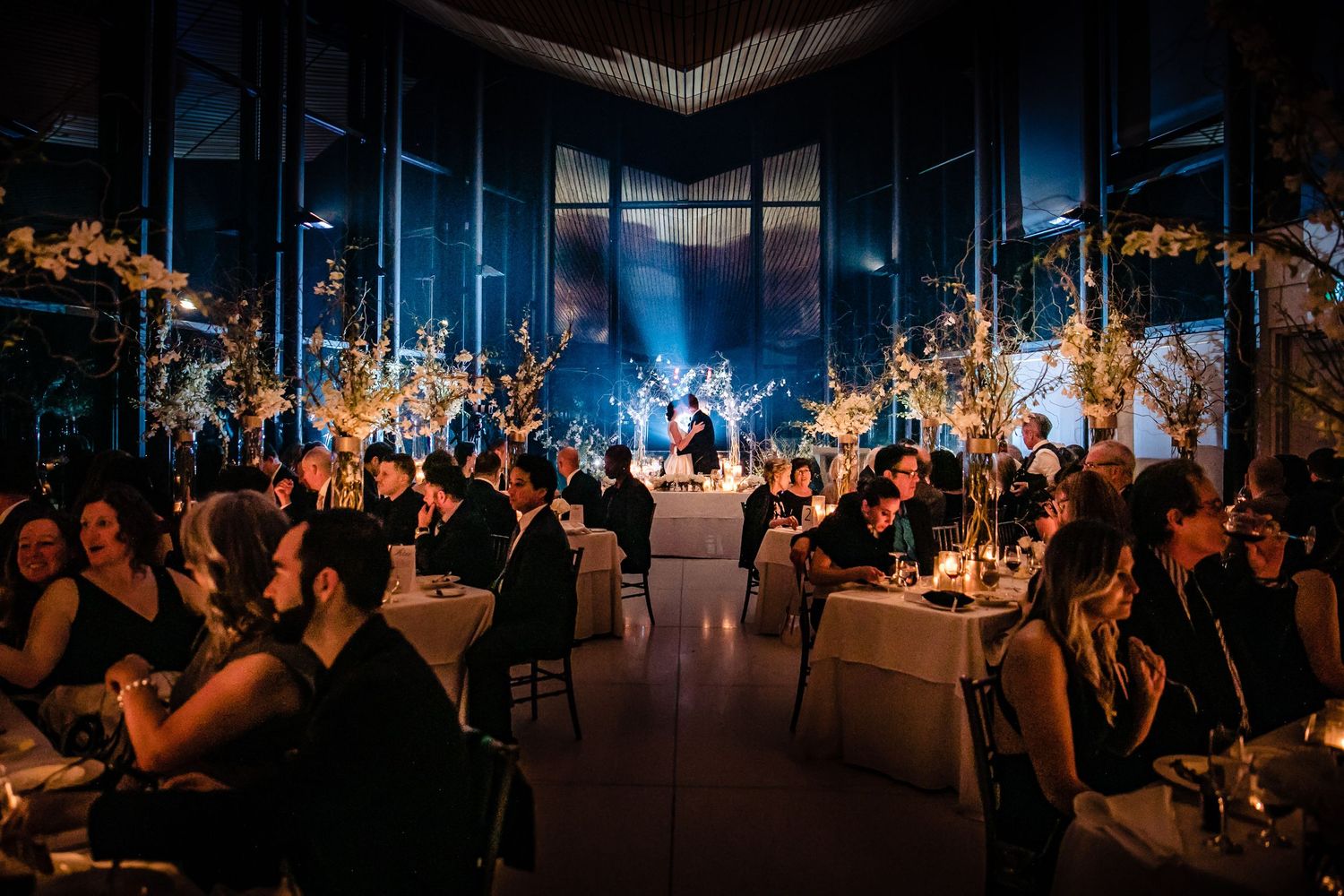 Elegant wedding reception venue with blue lighting and tables decorated with glowing orange floral arrangements.