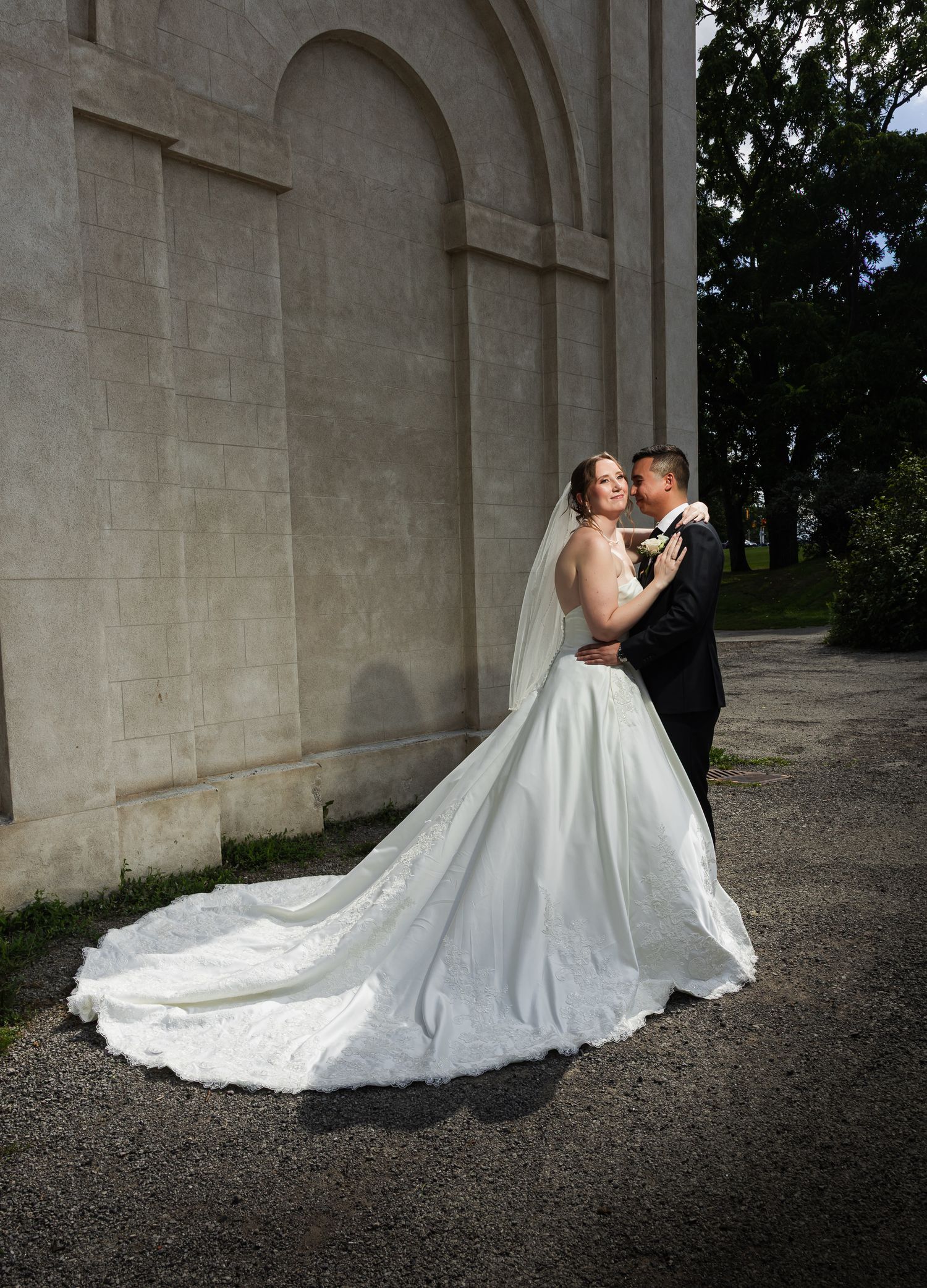 Bride in white ball gown with long train embraces groom in black tuxedo beside stone church wall.