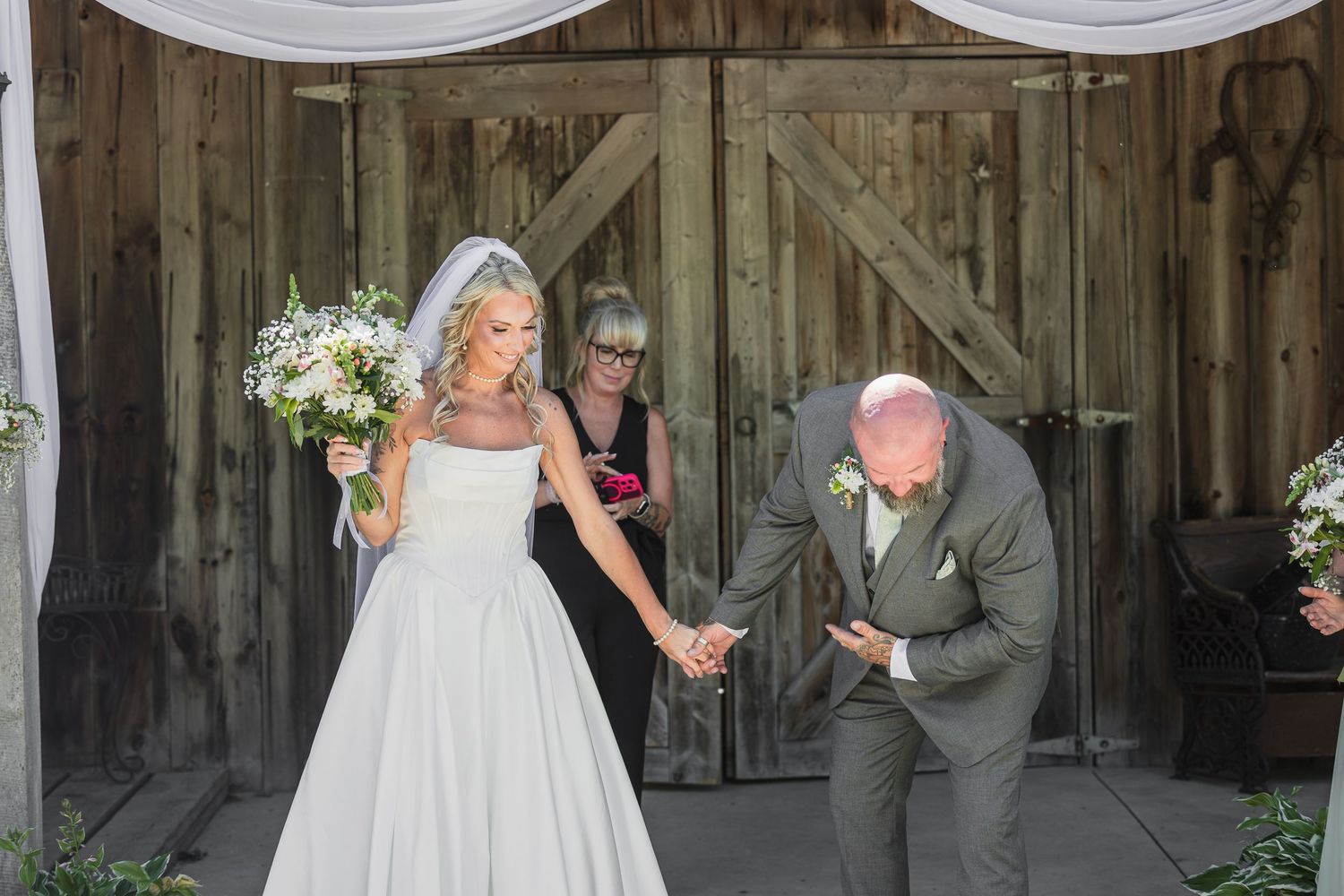 Bride and groom hold hands exiting barn wedding venue as officiant watches in background.