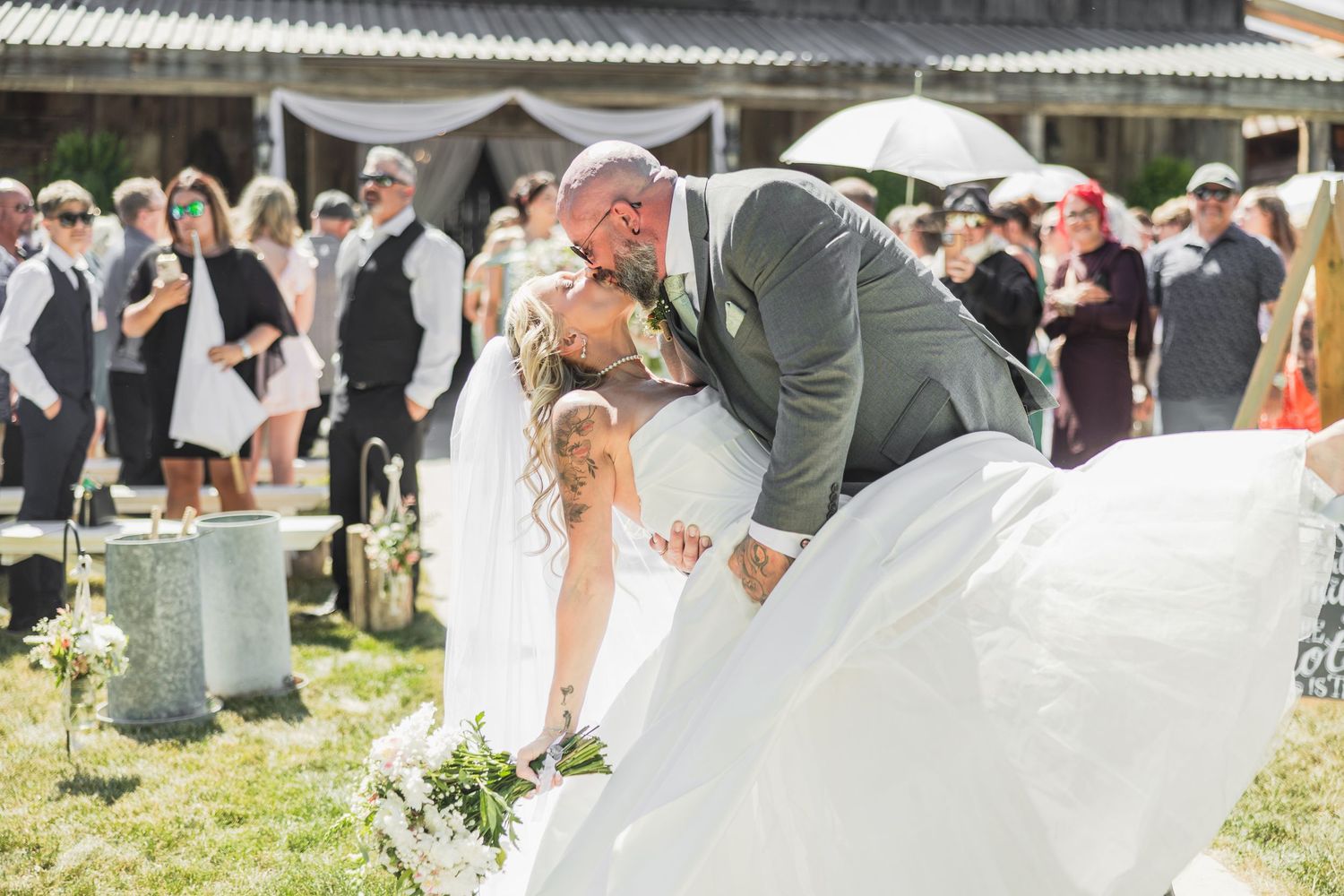 Bride and groom sharing a romantic dip kiss at outdoor wedding ceremony with guests watching in background.