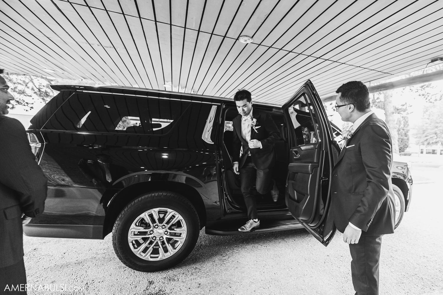 Wedding party members gather around black SUV under covered area.