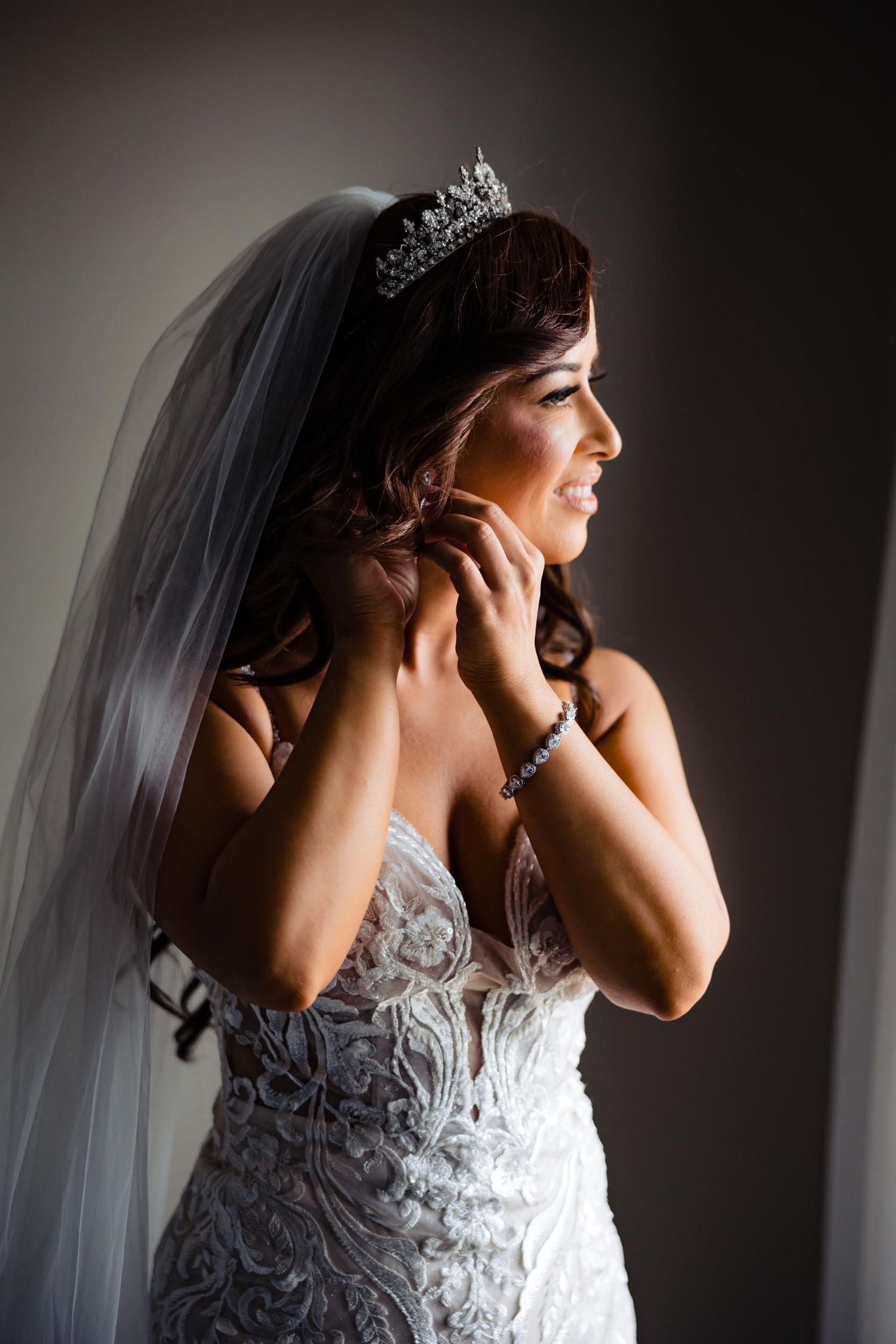 Side profile of a bride wearing a crystal crown and veil against dark background.