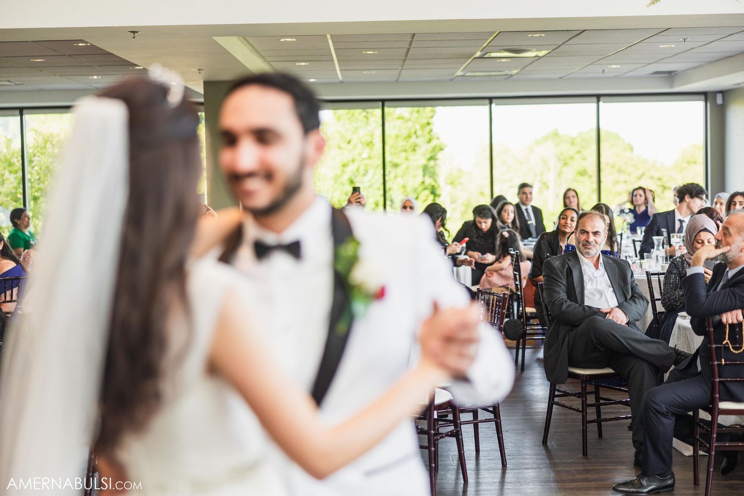 Wedding couple dances surrounded by guests in a bright reception venue with large windows.