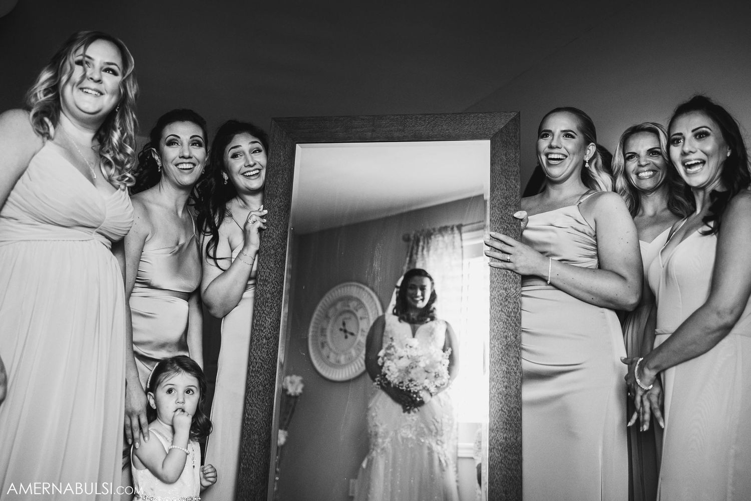 Bridesmaids in light colored dresses gather excitedly around doorway.