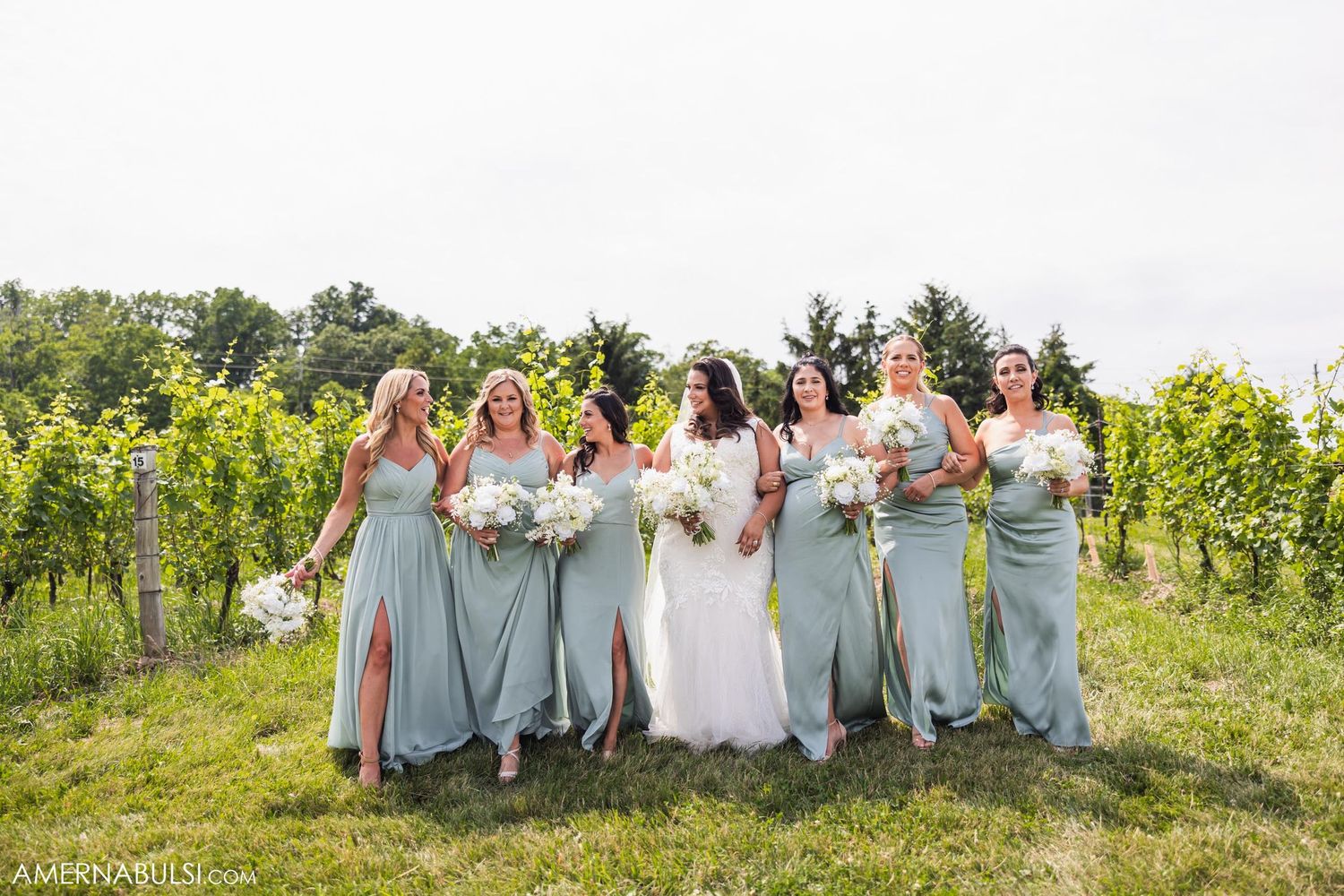 Bridesmaids in light blue dresses pose with bride among vineyard rows holding white bouquets.