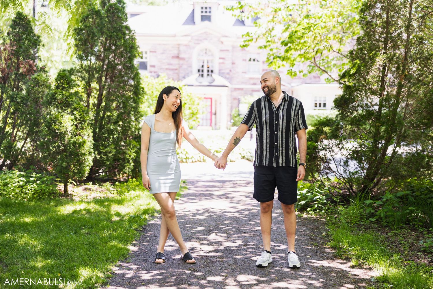 A couple holds hands while walking on a path with a mansion in the background.