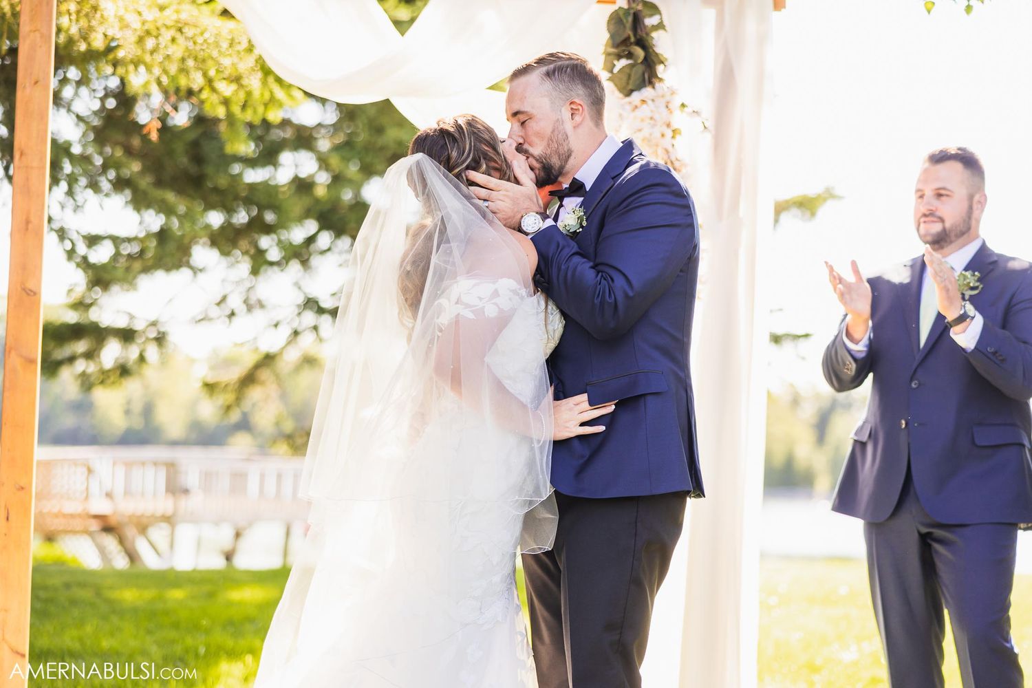 Newlyweds share their first kiss under a wooden arch as guests applaud at outdoor ceremony.