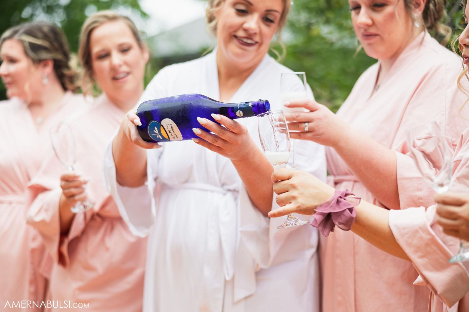 Group pouring champagne while wearing matching pink robes during pre-wedding celebrations.