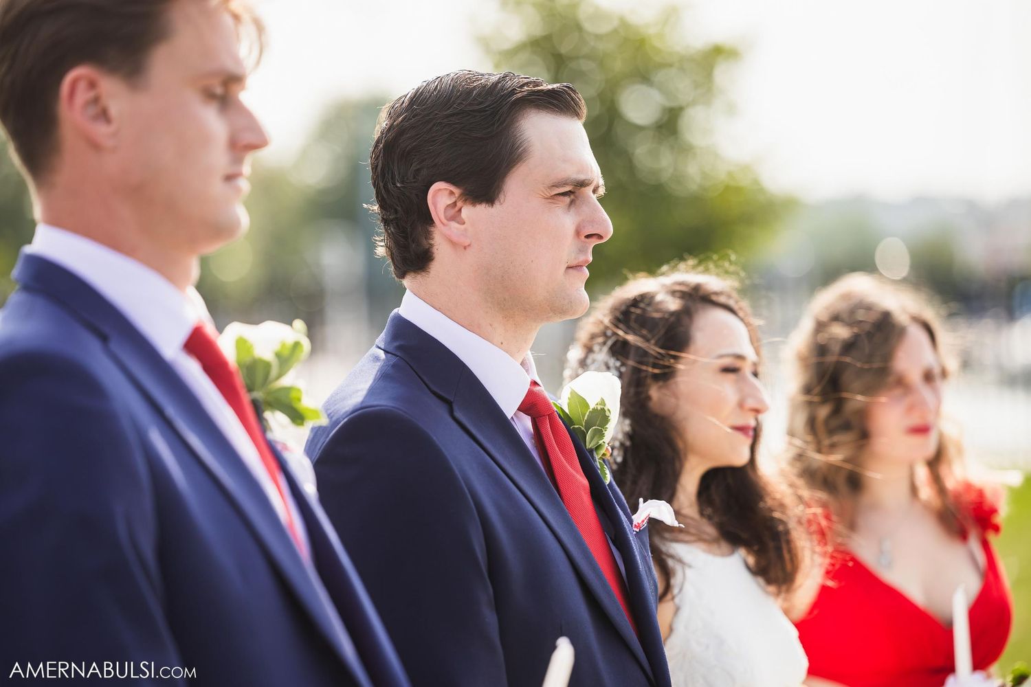 Wedding party members stand in a line wearing navy suits and red accents during the ceremony.