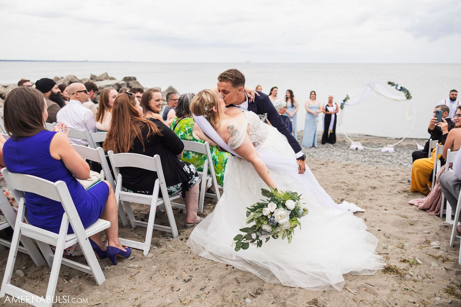 Beach wedding ceremony setup with white chairs and guests on the sandy shore.