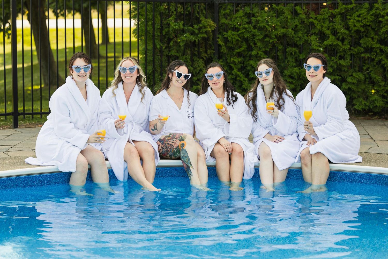 Group in white robes relaxing with drinks by pool during pre-wedding celebration.