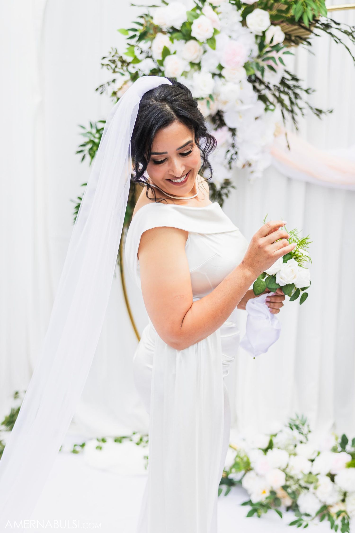 Bride in elegant white gown smiles while holding bouquet against floral backdrop.