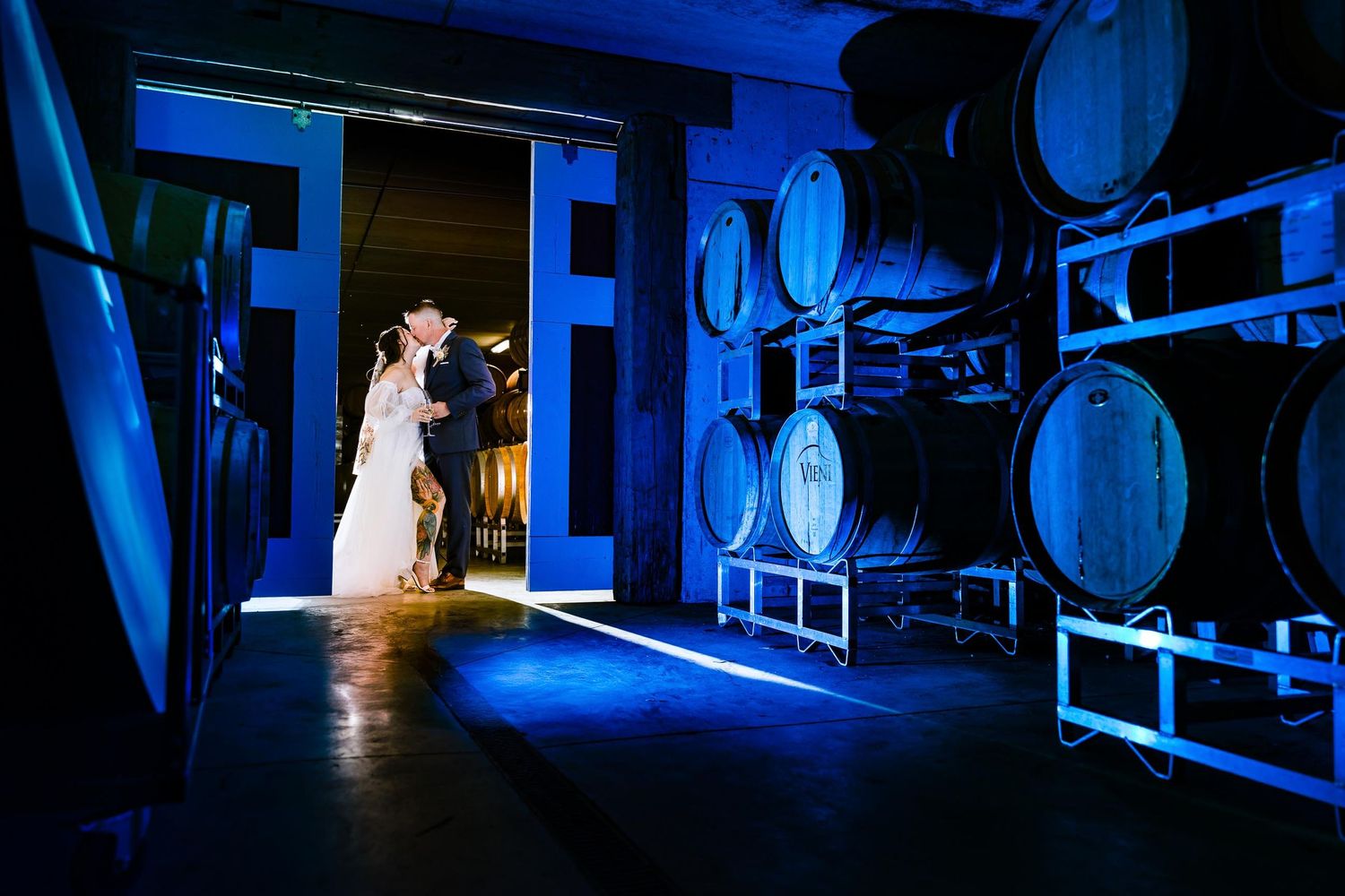 A couple stands in dramatic blue lighting among wine barrels in a cellar venue.