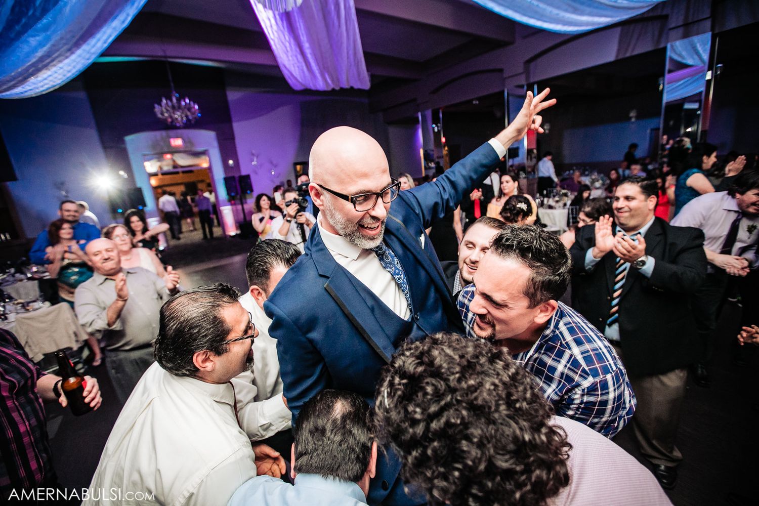 Wedding guests dance and celebrate on a crowded dance floor illuminated by purple and blue lighting.