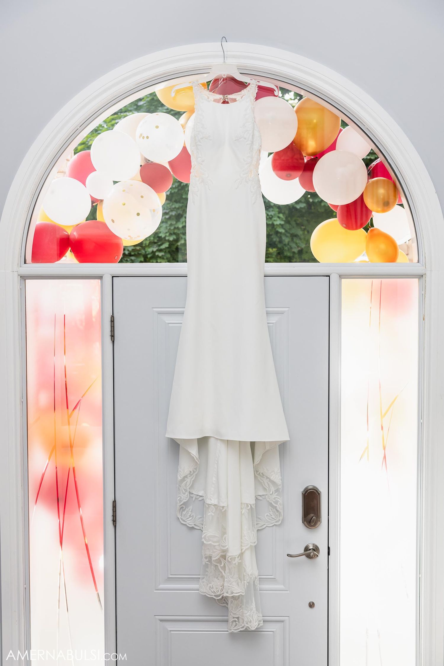White wedding dress hanging against arched doorway decorated with colorful balloons.