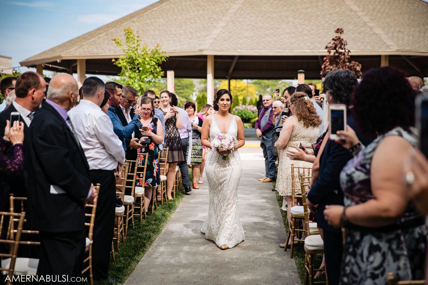 Outdoor wedding ceremony setup with guests seated under a beige tent canopy.